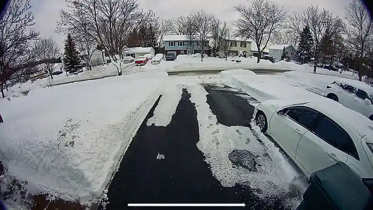 A fisheye-lens security camera view of a snow-covered residential driveway with cleared paths, showing footprints in the…