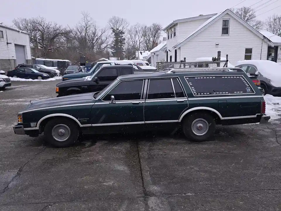 A dark green 1980s Ford station wagon is parked in a snowy lot, its rear window covered with a sticker map of all 50 U.S…