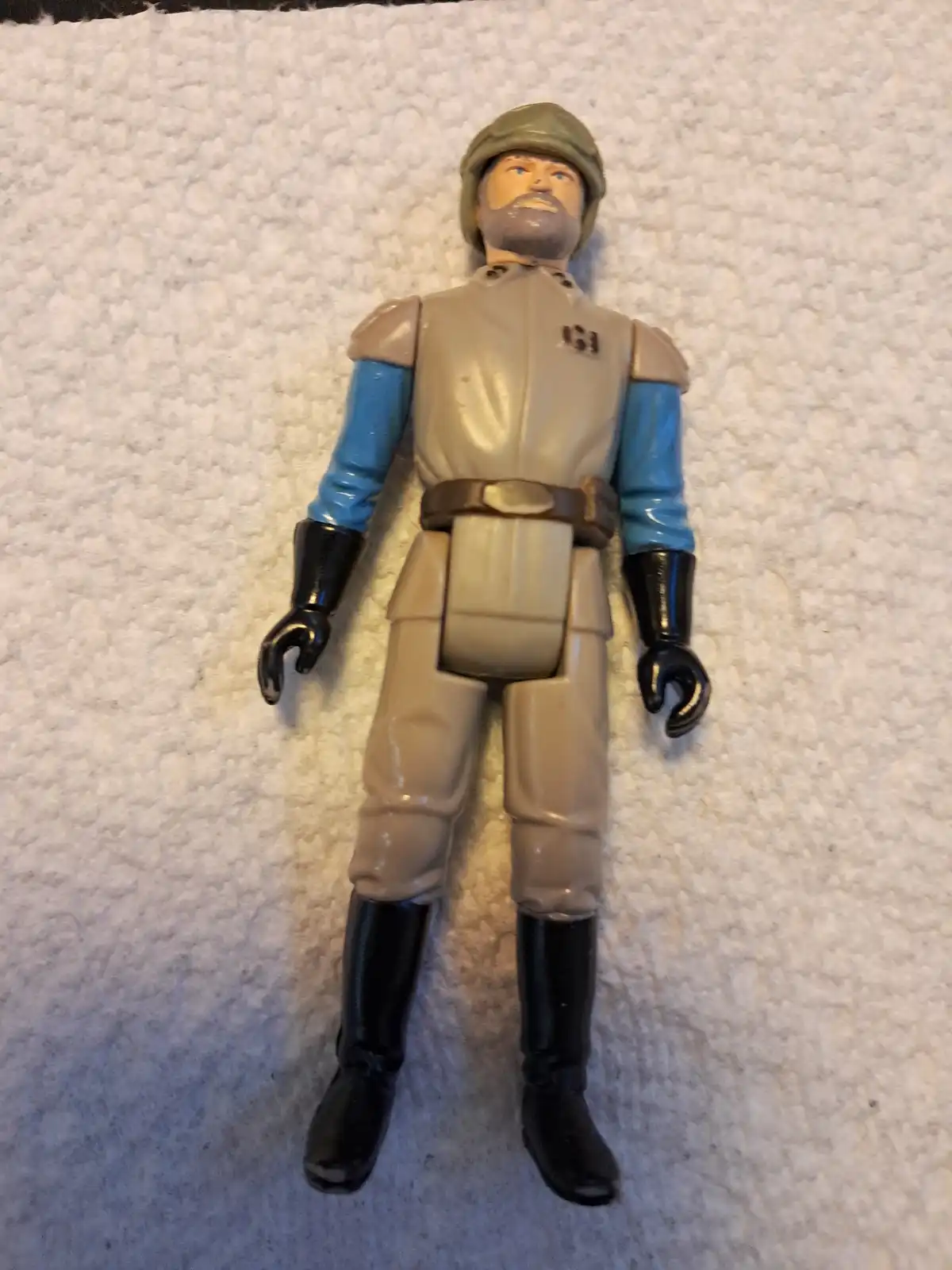 A vintage Star Wars action figure, resembling a Rebel officer with a beard, helmet, and tan-and-blue uniform, lies flat on a…