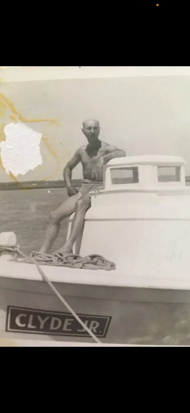 A vintage black-and-white photo shows a shirtless, bald man sitting on the bow of a small boat named 'CLYDE JR.' with water…
