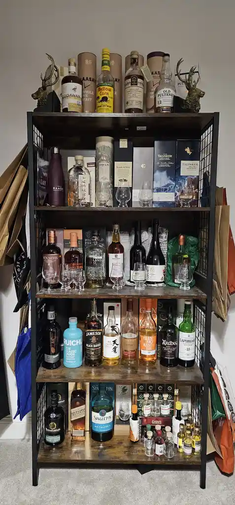 A tall, dark metal shelving unit with wooden shelves is filled with numerous bottles of whiskey, whisky, and related…