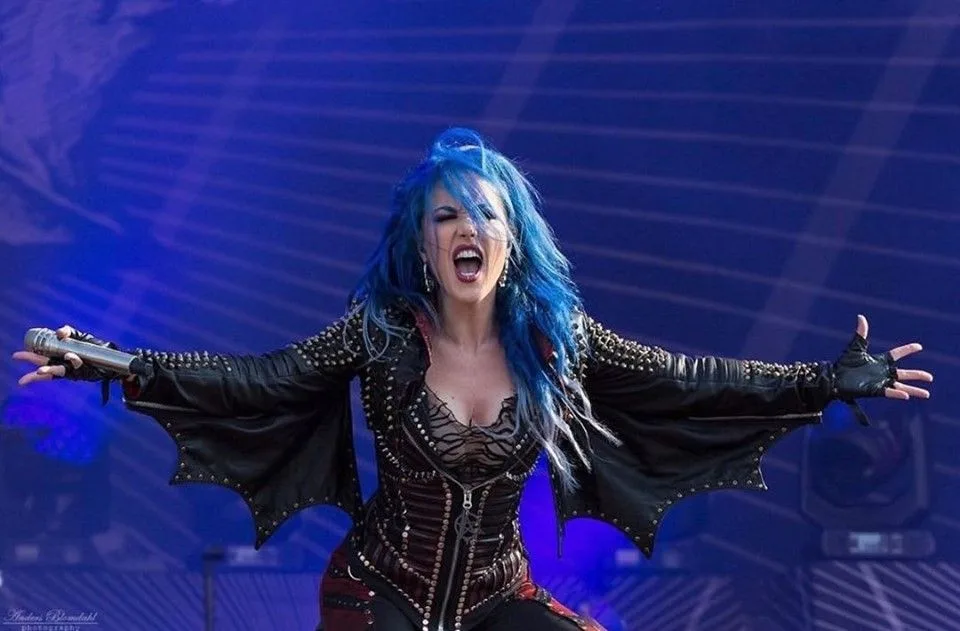 A female singer with vibrant blue hair and dramatic stage makeup is captured mid-performance, arms wide open, holding a…