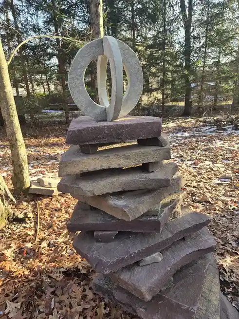 A tall, precarious stack of flat, rough-hewn stones sits outdoors in a wooded area with fallen leaves and patches of snow on…