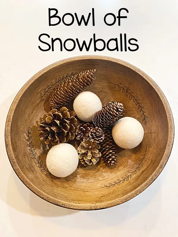 A wooden bowl with decorative leaf etchings contains several pinecones and three white, round, felt-like objects resembling…