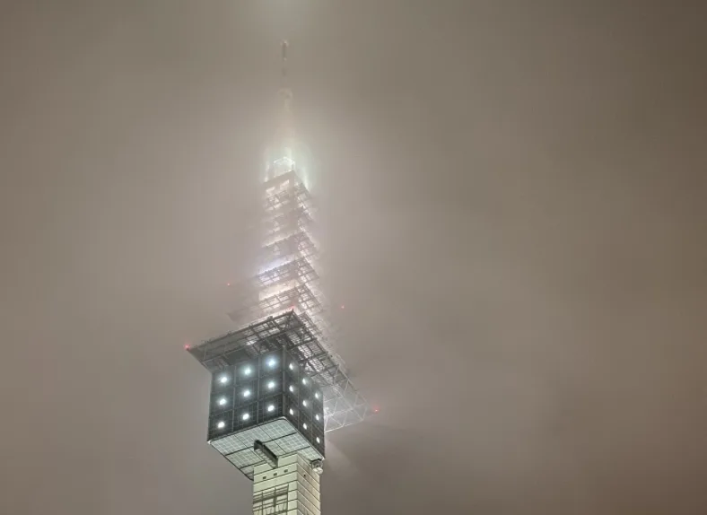 a tall, illuminated tower is partially obscured by thick fog, with only its lower section and bright lights visible against…