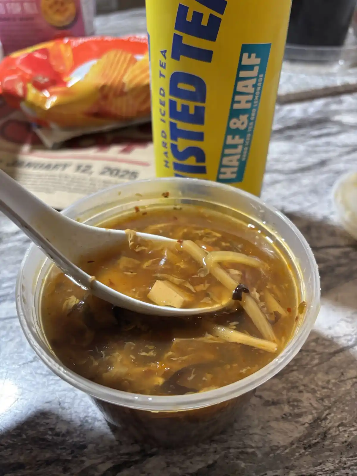 A close-up photo shows a clear plastic cup of hot, thick, brownish soup with tofu and noodles, a white spoon resting in it…