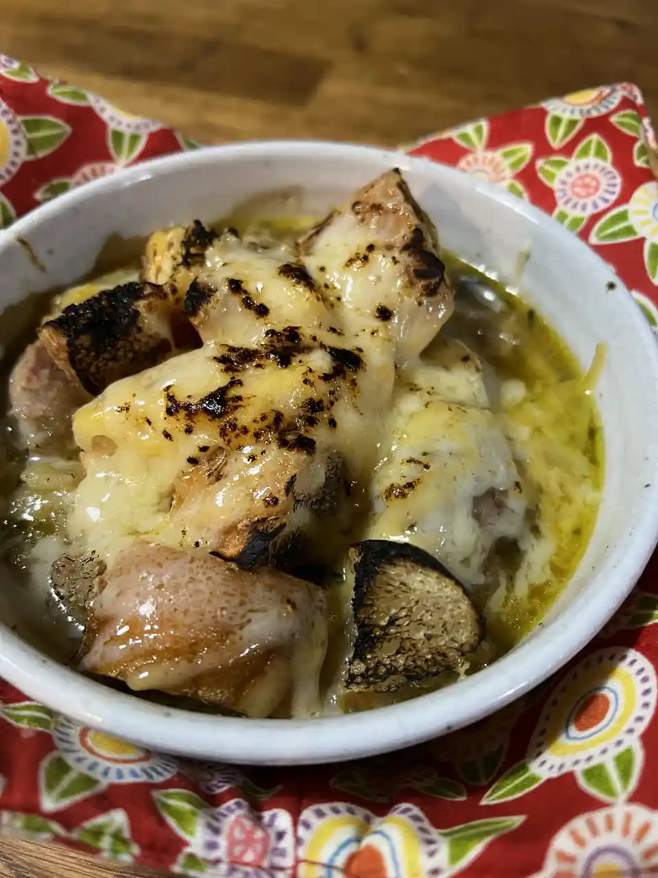 A close-up photo shows a white bowl filled with a cheesy, bubbling dish that appears to be French onion soup, but the cheese…