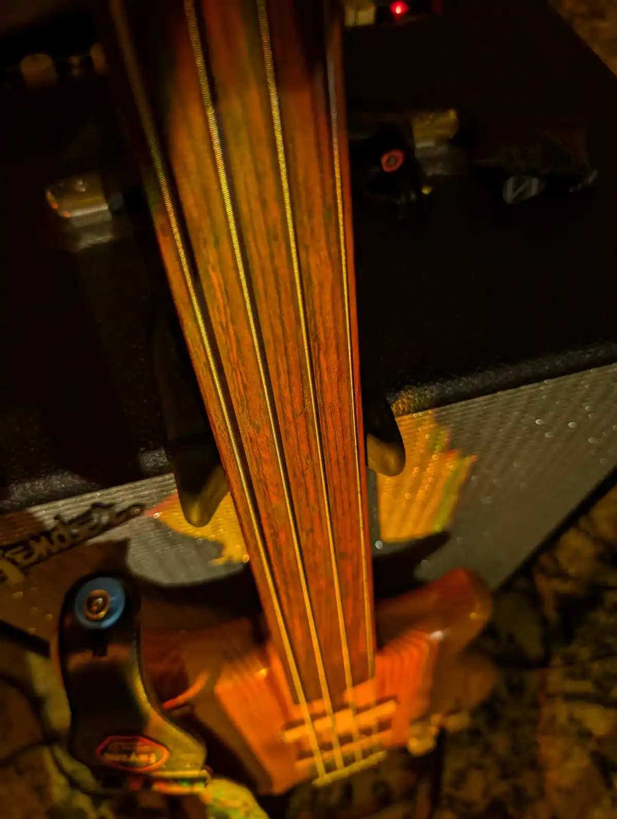 A close-up, dimly lit photo shows the neck and strings of a wooden bass guitar leaning against a black amplifier. The bass…