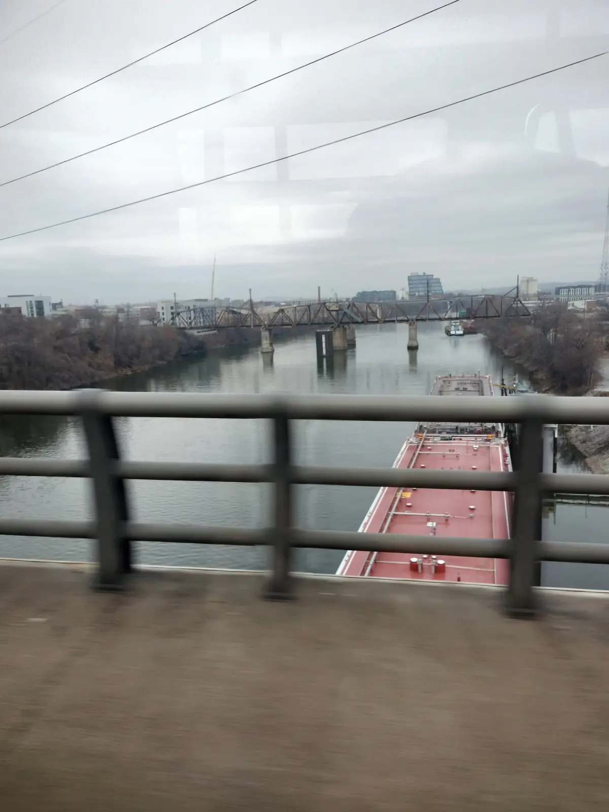 A blurry photo taken from a moving train window shows a red barge on a river below, with a metal truss bridge in the…