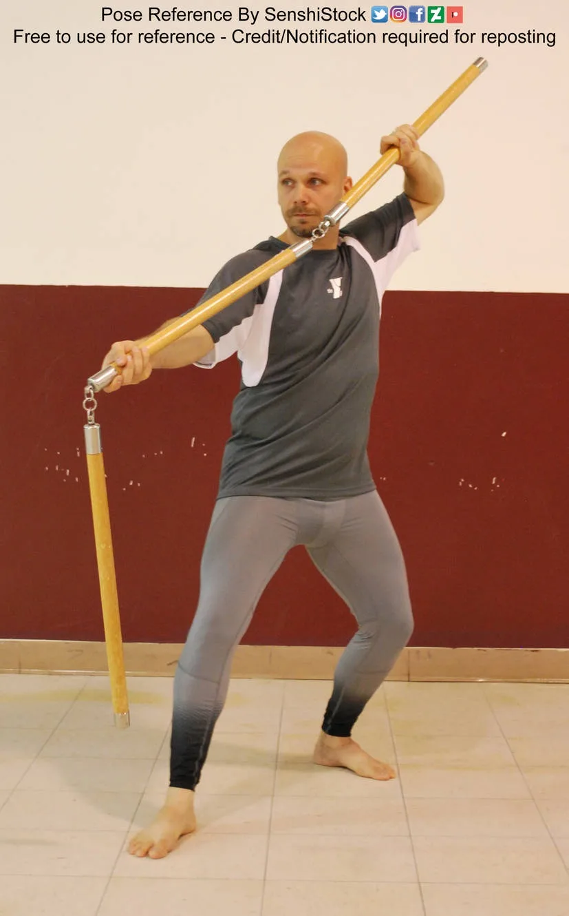 A bald man in a grey and white shirt and grey leggings stands barefoot on a tiled floor, holding a long wooden nunchaku in a…