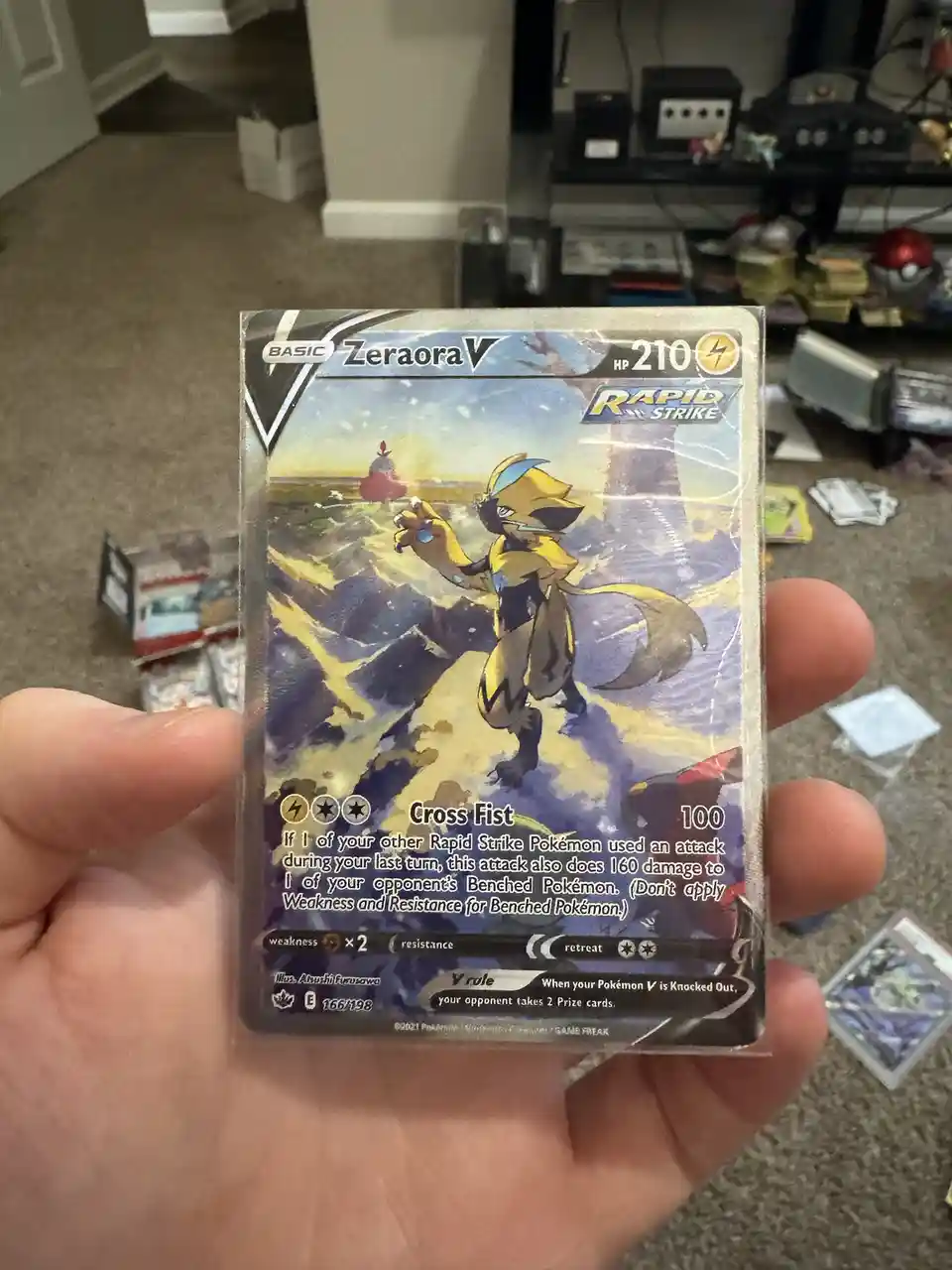 A hand holds a heavily scratched and damaged Pokémon TCG card of Zeraora V, with visible wear on its edges and surface. The…