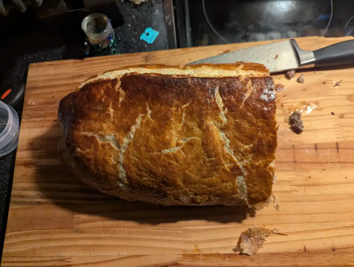 A loaf of golden-brown bread with a cracked crust sits on a wooden cutting board, with a large kitchen knife resting beside…