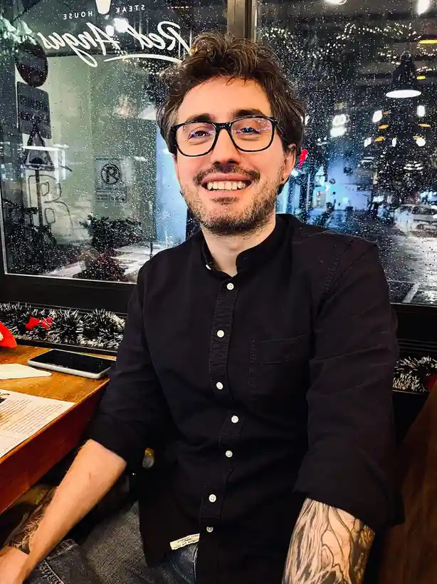 A man with dark hair, glasses, and a beard smiles at the camera while sitting inside a restaurant. Raindrops streak the…