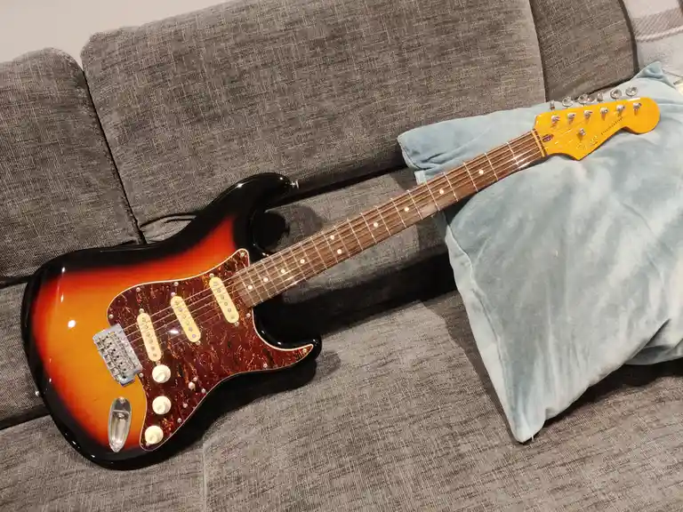 A sunburst-finish Squier Stratocaster electric guitar rests diagonally across a grey fabric couch, its headstock resting on…