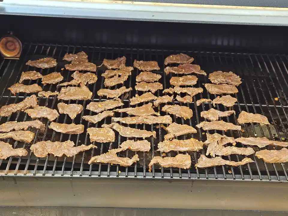 A close-up photo shows numerous thin, seasoned meat strips sizzling on a black metal grill grate inside a barbecue grill. A…