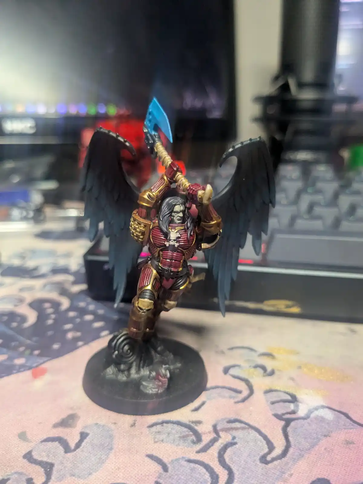 A painted miniature of a winged warrior in red and gold armor, holding a blue axe, stands on a base. The image is overlaid…