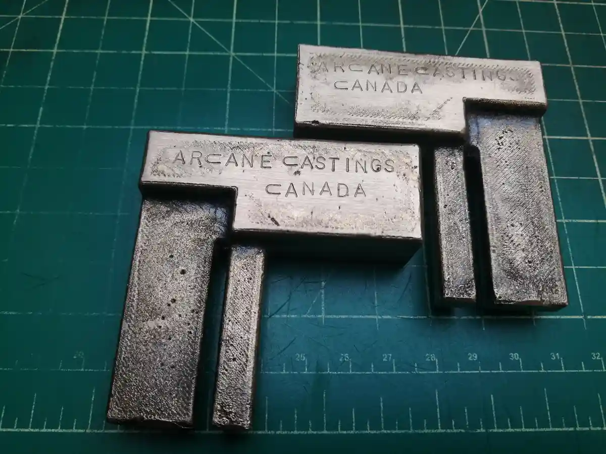 Two metallic, T-shaped castings with 'ARCANE CASTINGS CANADA' engraved on their flat tops sit on a green cutting mat with…