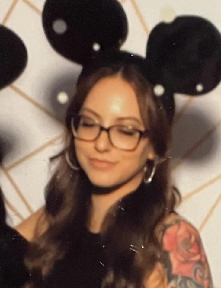 A woman with long brown hair, glasses, and a visible red rose tattoo on her right shoulder wears black Mickey Mouse ears…