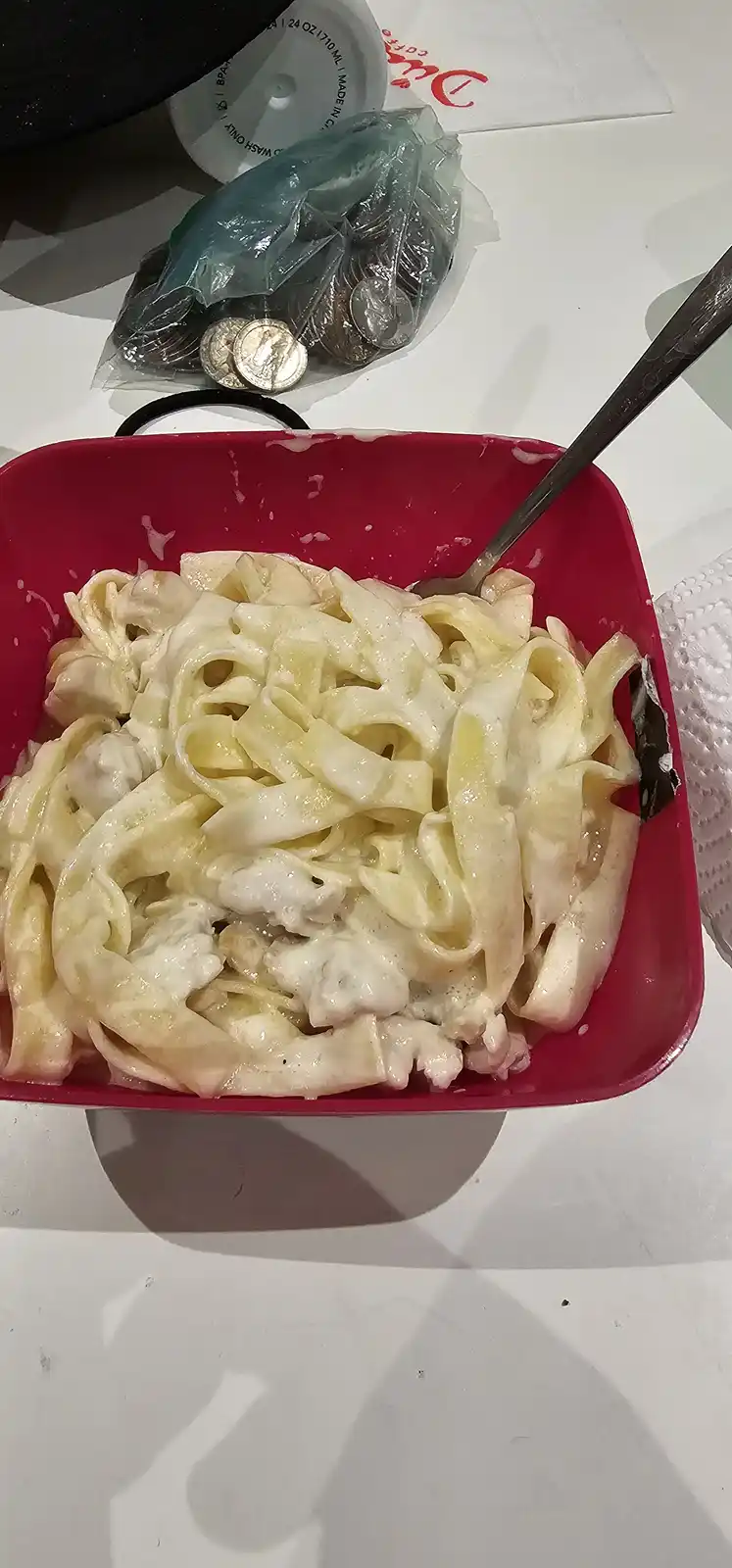 A red plastic bowl filled with creamy fettuccine pasta sits on a white table, with a metal fork inserted. Behind it, a clear…
