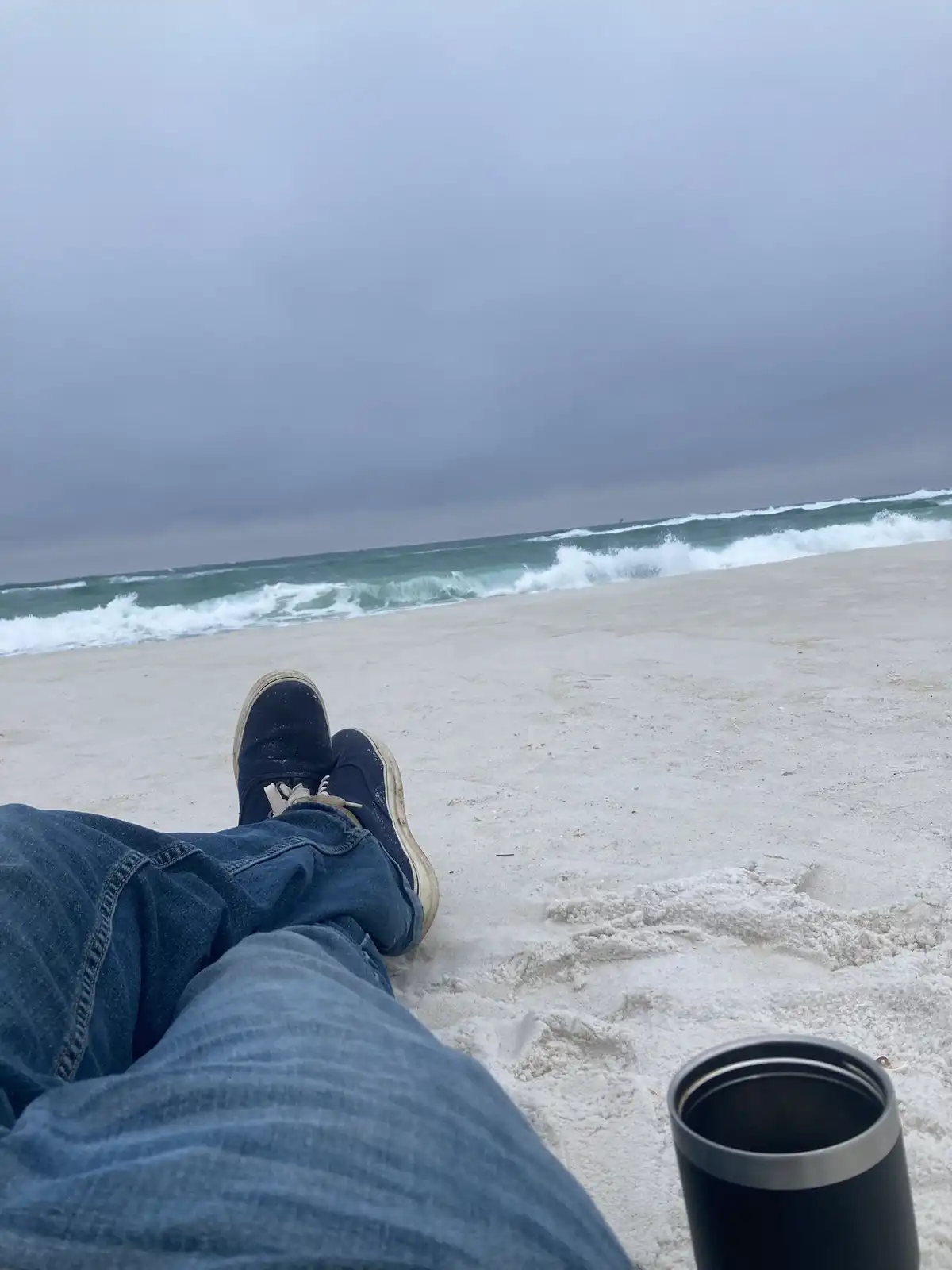A first-person view of someone sitting on a sandy beach with their legs and blue jeans visible, wearing dark sneakers. The…