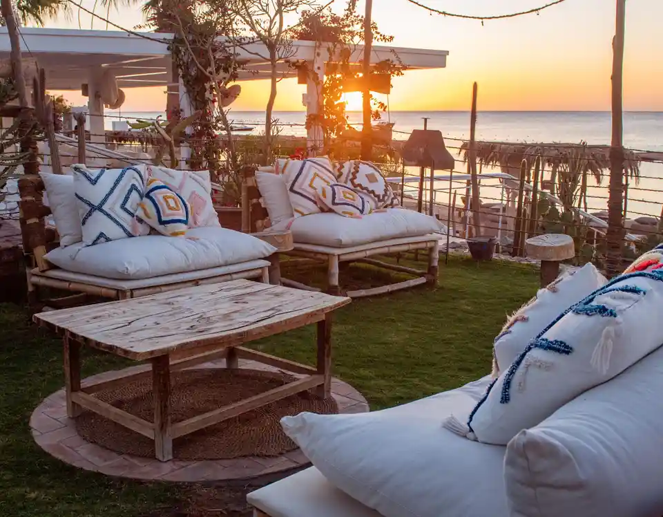A serene outdoor patio at sunset features bamboo furniture with white cushions and colorful patterned pillows, overlooking…