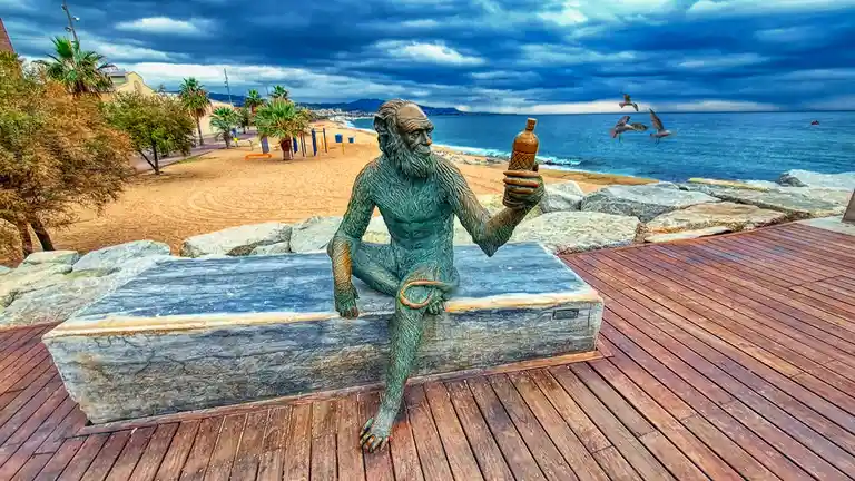 A bronze statue of a bearded, ape-like humanoid sits on a stone slab at a seaside promenade, holding a bottle of alcohol…