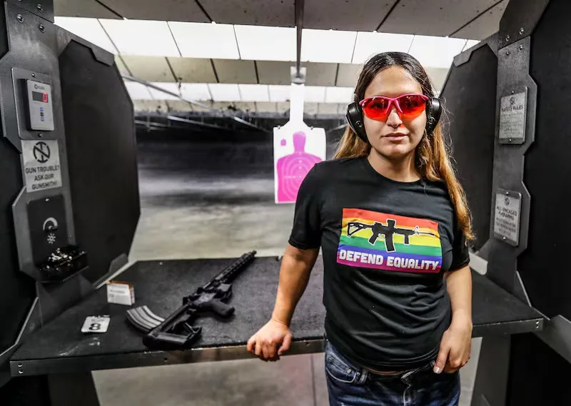A woman stands in a shooting range wearing red-tinted safety glasses and ear protection, posing confidently in front of a…