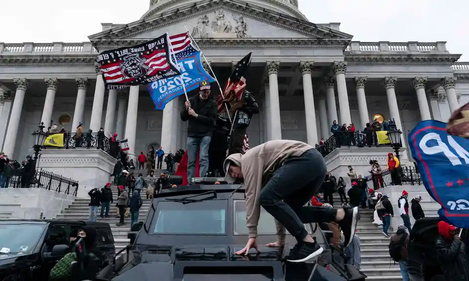 A person in a beige hoodie climbs onto the roof of a black armored vehicle in front of the U.S. Capitol building, while…