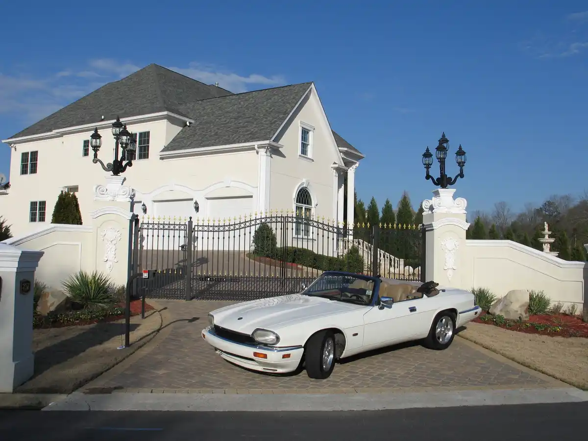 A white Jaguar XJS convertible is parked on a paved driveway in front of an ornate, gated mansion with white pillars, black…