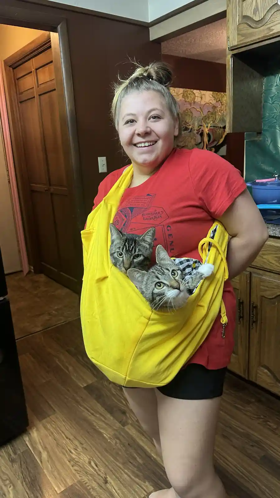 A smiling woman in a red shirt and black shorts stands indoors, wearing a bright yellow baby carrier sling that holds two…