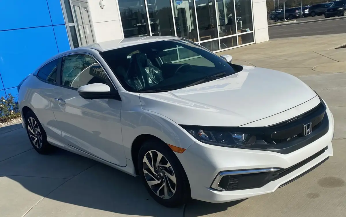 A white Honda Civic coupe is parked outside a dealership with a blue wall and glass windows in the background. The car has…