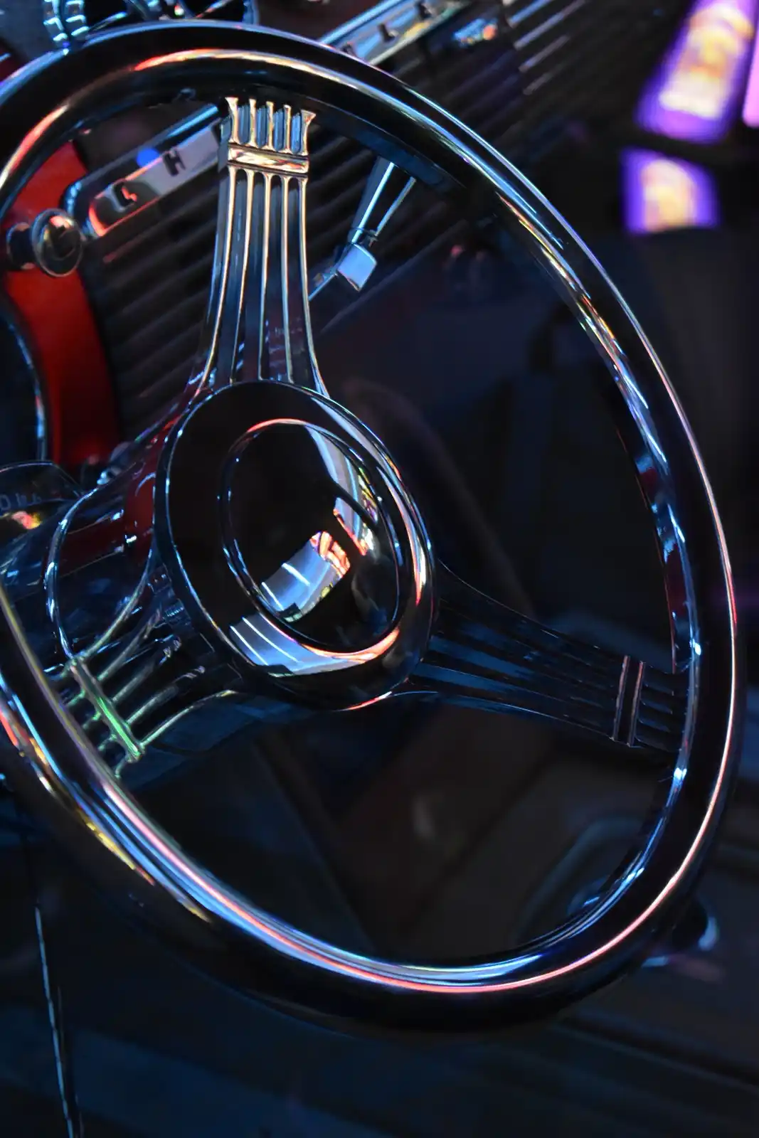 A close-up photo of a shiny, chrome classic car steering wheel with a three-spoke design, reflecting colorful neon lights…