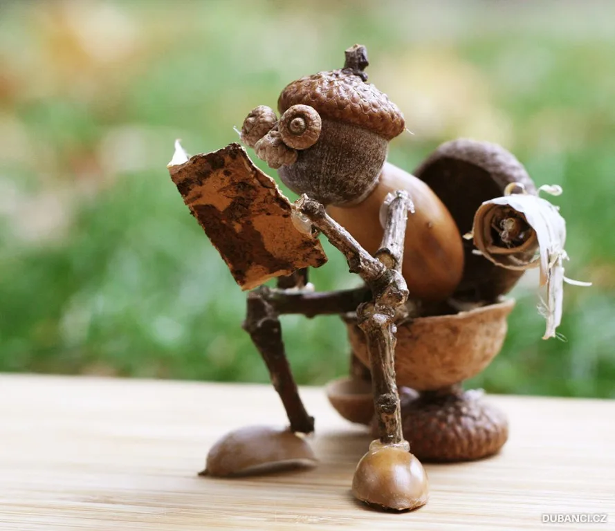A whimsical sculpture made of acorns and twigs depicts a tiny figure sitting on a wooden surface, holding a small book open…