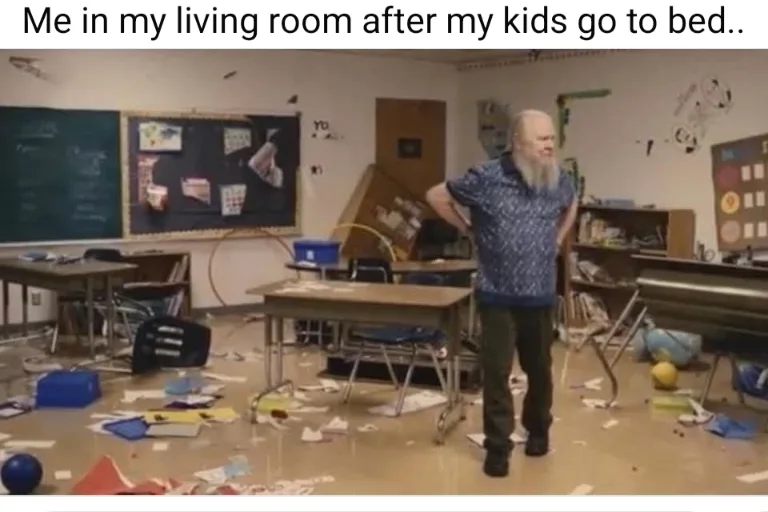 A man with a long white beard and blue patterned shirt stands in a classroom with desks, chairs, and papers scattered…