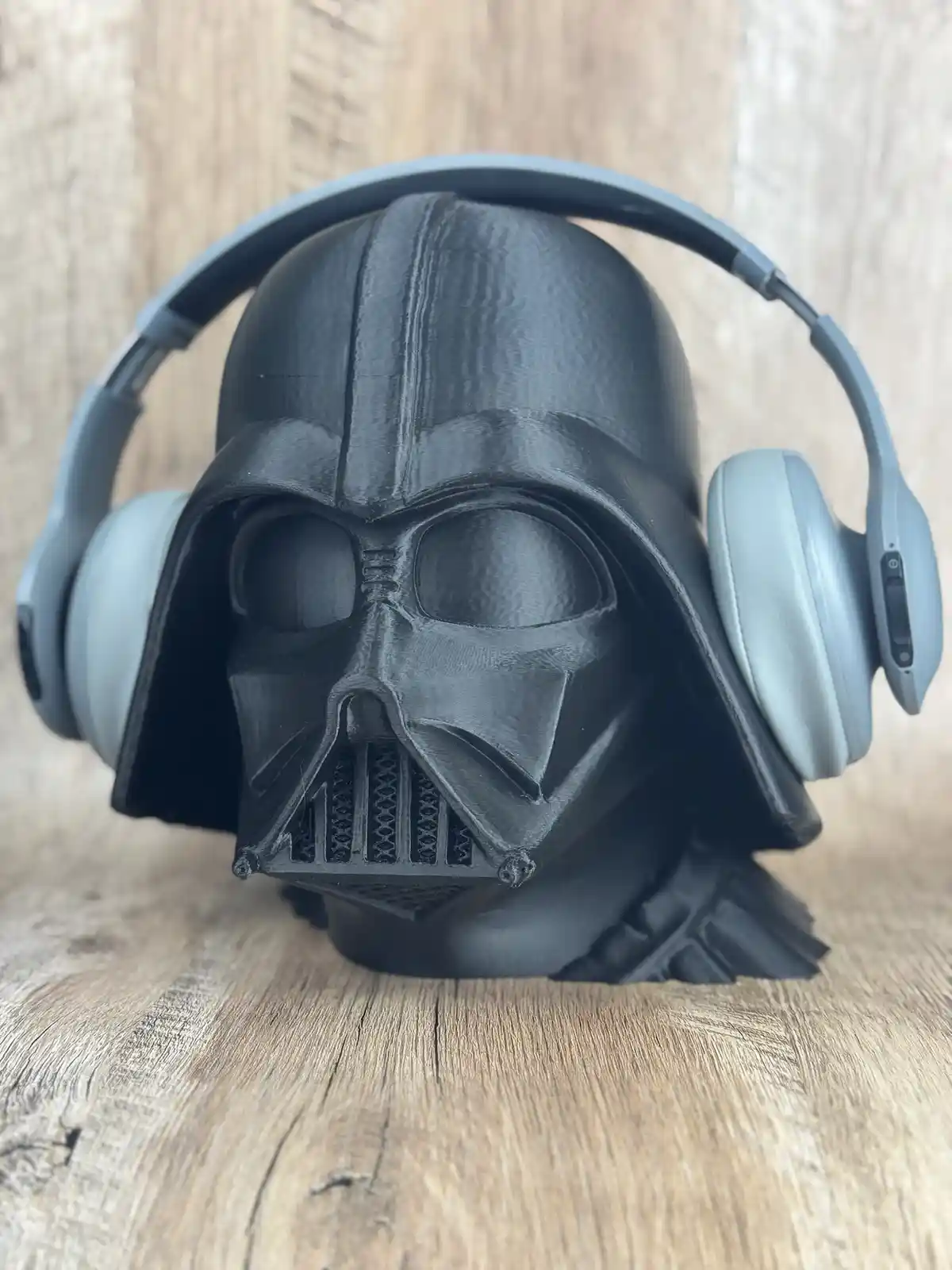 A 3D-printed Darth Vader helmet head sculpture sits on a wooden surface, with a pair of light gray over-ear headphones…