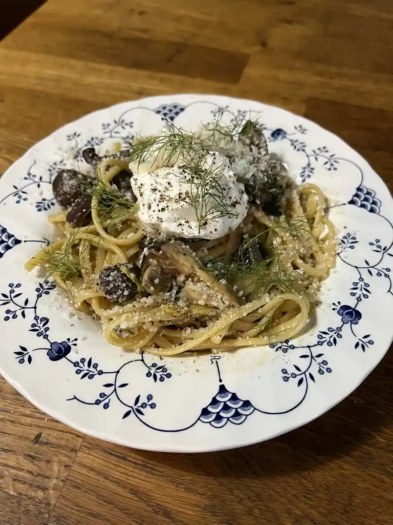 A close-up photo of a plate of pasta with mushrooms, grated cheese, dill, and a dollop of cream on top, served on a white…