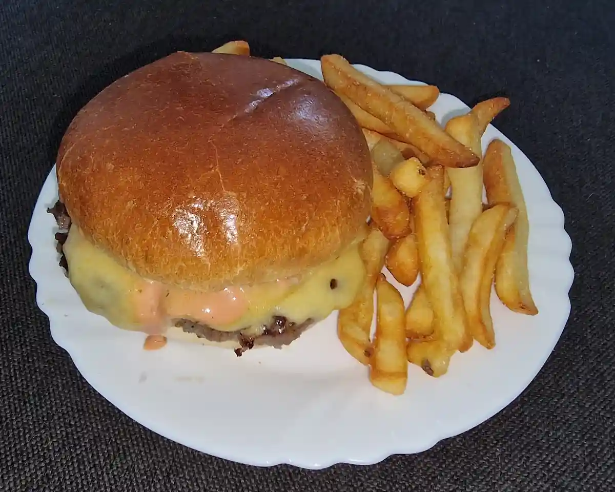 A close-up photo of a cheeseburger with melted cheese and sauce oozing out, served with french fries on a white plate…
