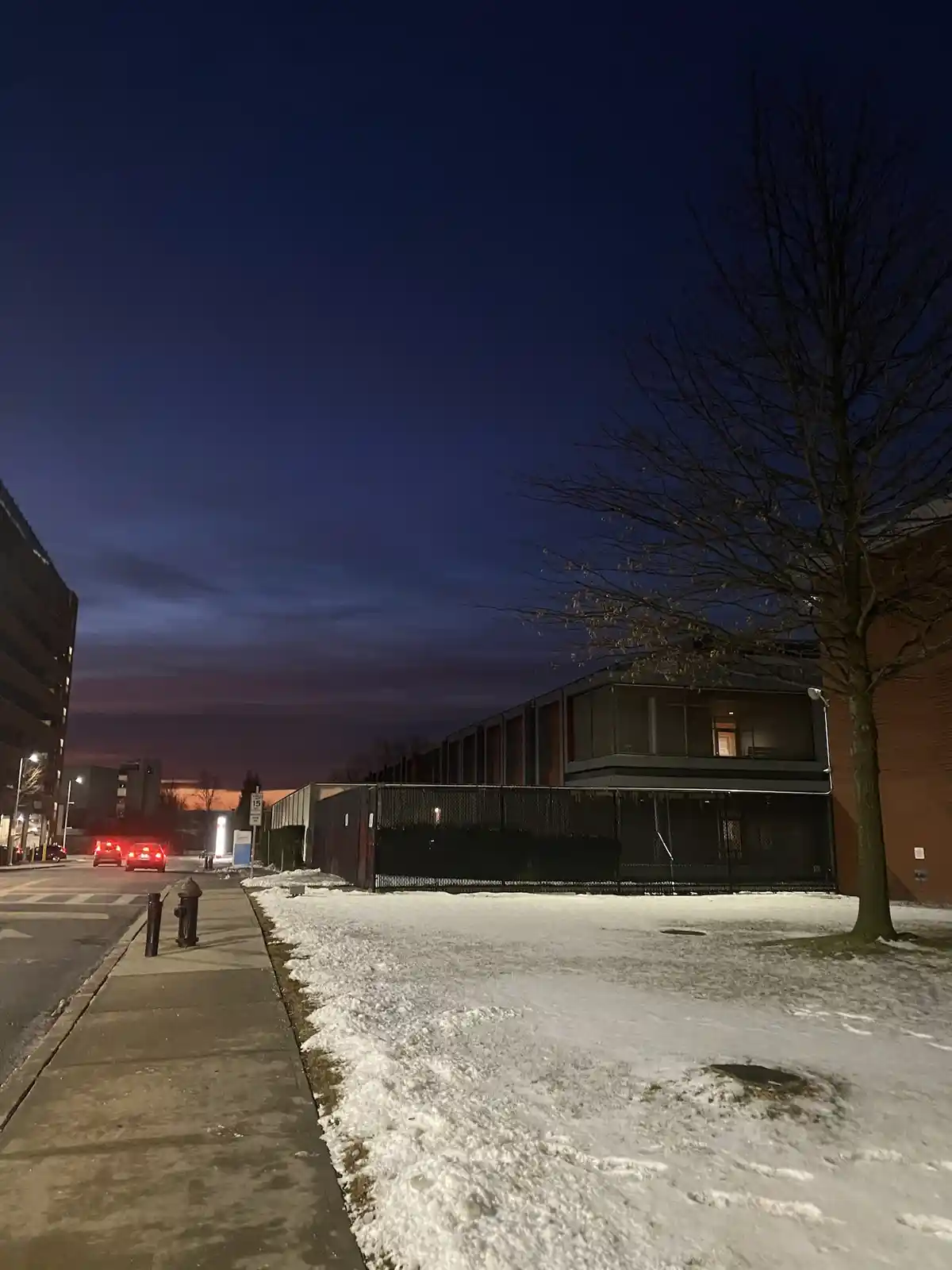A nighttime street scene with snow on the ground, a sidewalk, a fire hydrant, and buildings under a dark blue sky with a…