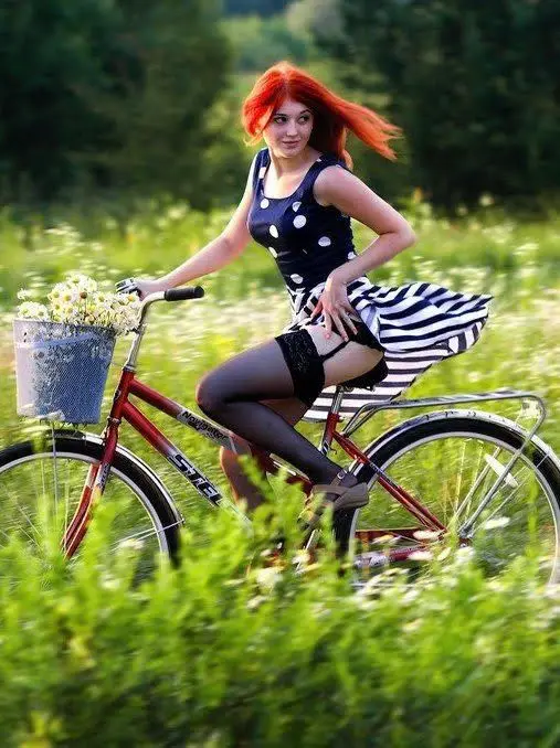 A woman with bright red hair is posing on a red bicycle in a field of tall green grass and white wildflowers. She wears a…