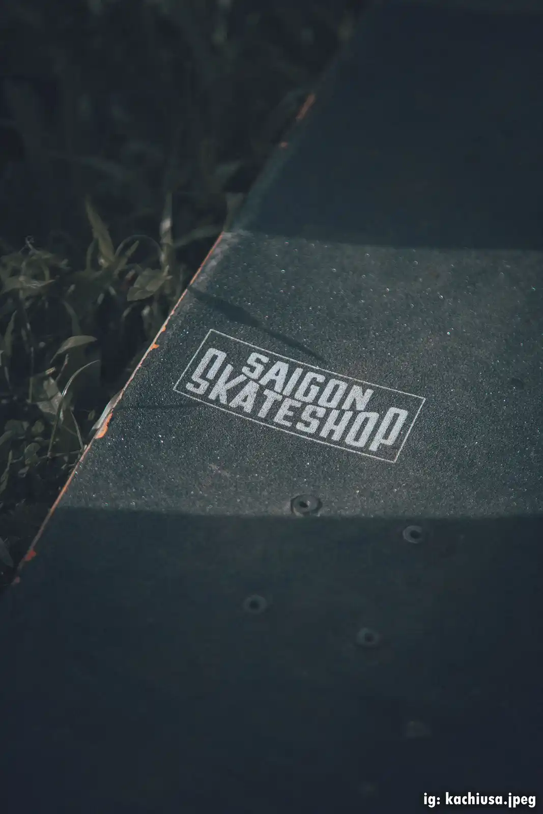 A close-up photo of a dark skateboard deck resting on grass, showing a white rectangular sticker that reads 'SAIGON…