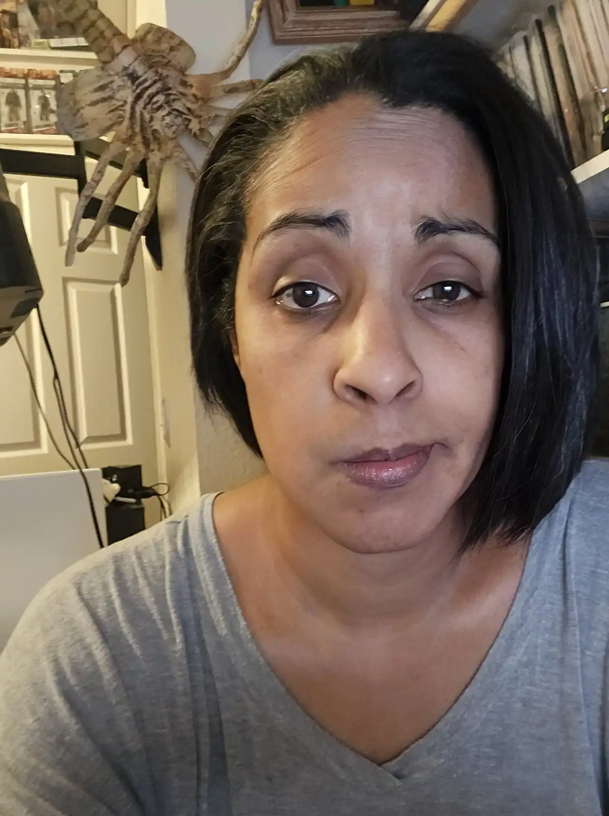 A woman with short dark hair and a gray shirt stares directly at the camera with a deadpan, unimpressed expression. Behind…