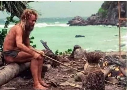 A man with long hair and beard, wearing minimal clothing, sits on a log near a campfire on a tropical beach, holding a…