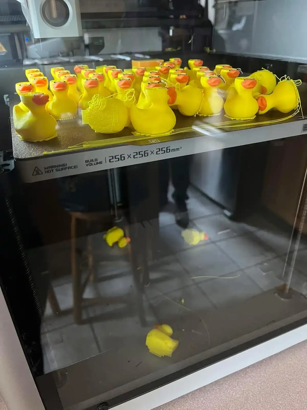 An image shows the inside of a 3D printer with dozens of yellow rubber duck figurines printed on the build plate, some still…