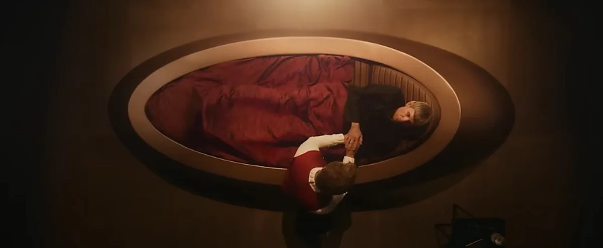 An overhead shot shows a person in a red and white outfit kneeling beside an oval bed where another person lies under a red…