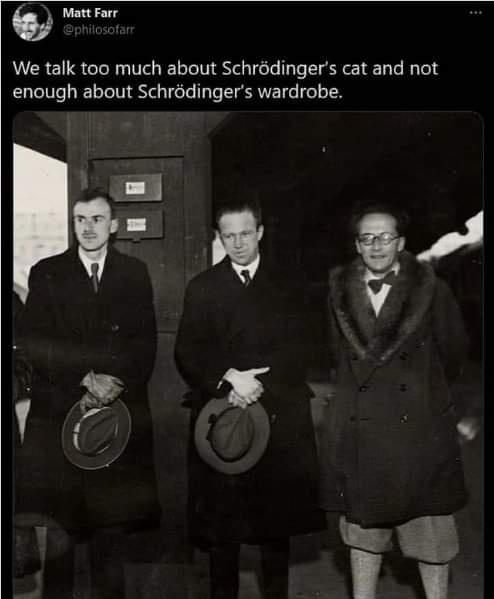 A black-and-white photo shows three men in coats standing outdoors. The man on the right wears a fur-collared coat over…
