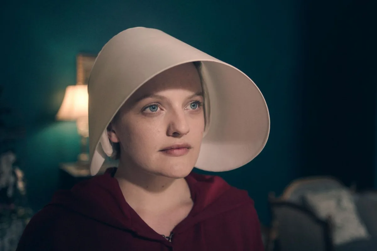 A woman wearing a white wide-brimmed hat and a red hooded garment looks directly at the camera with a serious expression in…