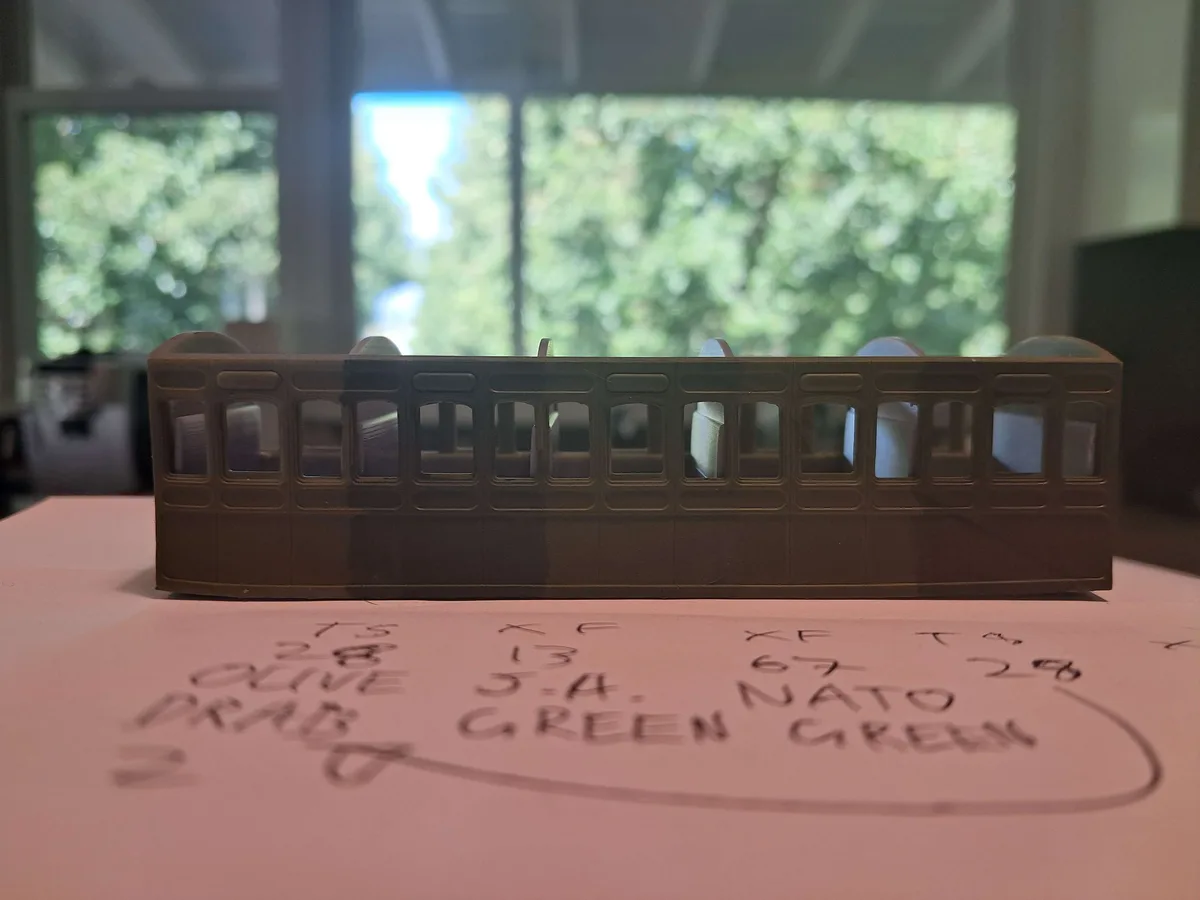 A small, brown model train car sits on a table with handwritten notes nearby listing military green color codes like 'OLIVE…