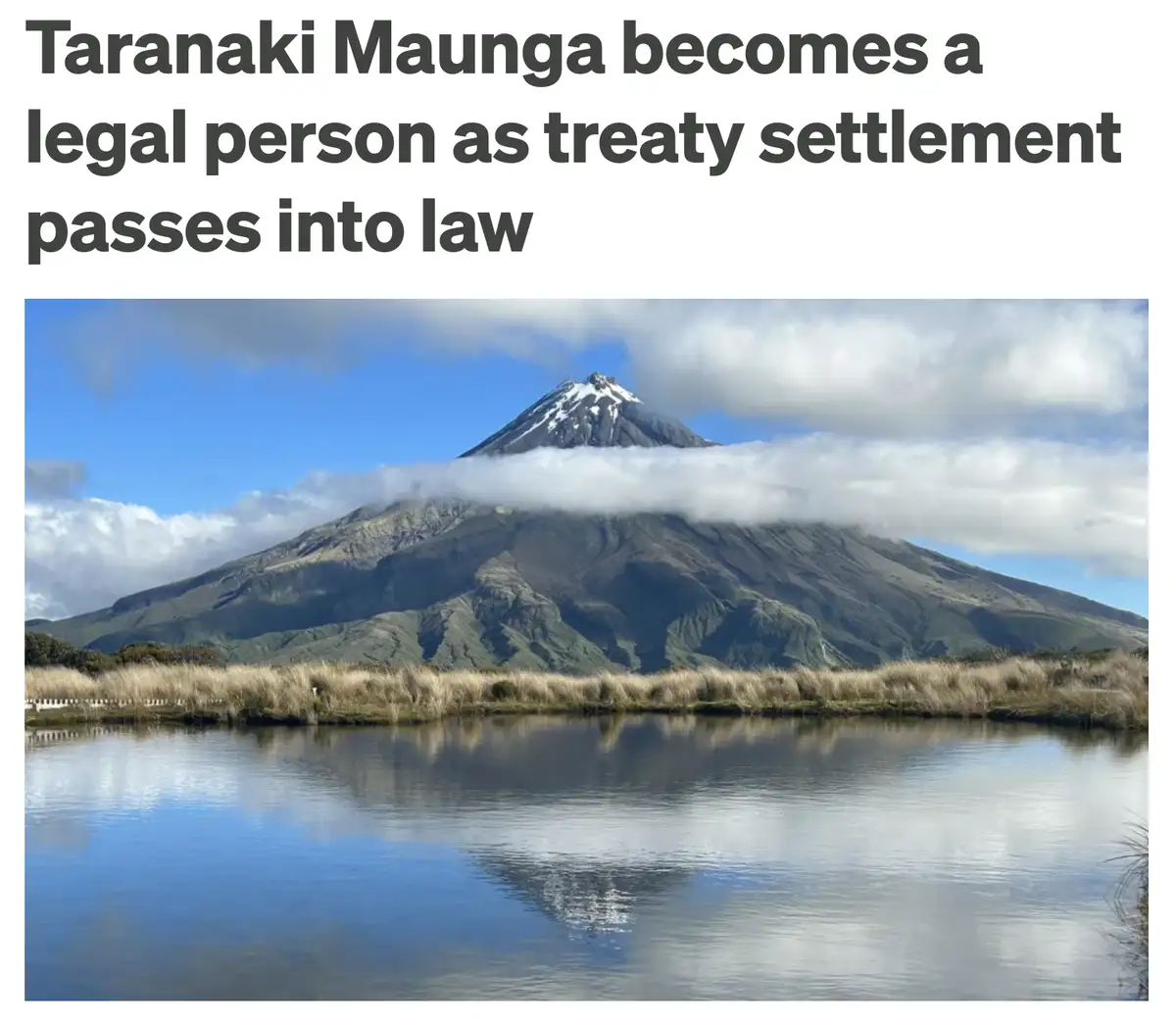 A scenic photograph shows Taranaki Maunga, a snow-capped mountain partially obscured by clouds, reflected in a calm body of…