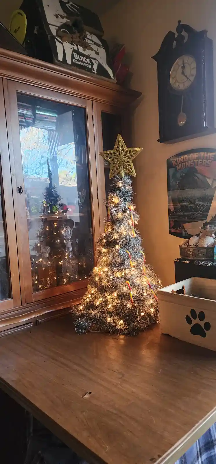 A small, silver tinsel Christmas tree with warm lights and a gold star topper sits on a wooden counter. Behind it is a…