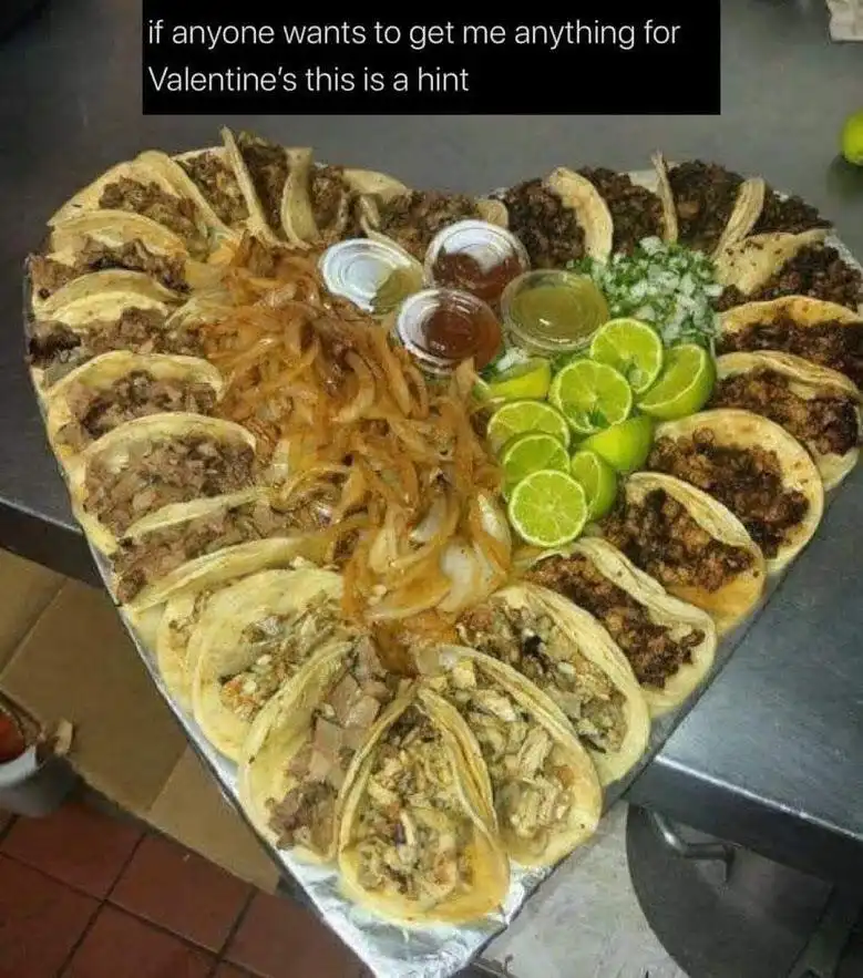 A large heart-shaped platter filled with numerous tacos, garnished with sliced limes, chopped onions, and small jars of…